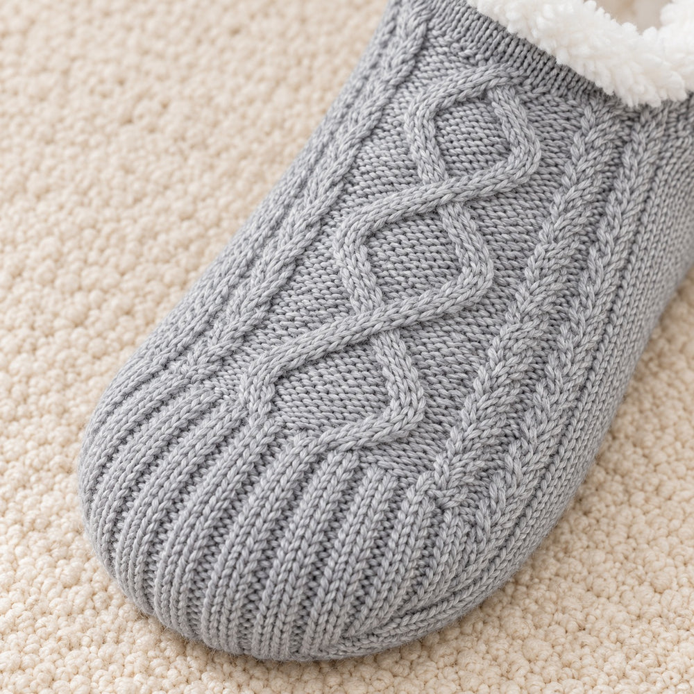 Nahaufnahme des grauen Zopf- und Rautenmusters einer Thermosocke auf einem cremefarbenen Teppich.