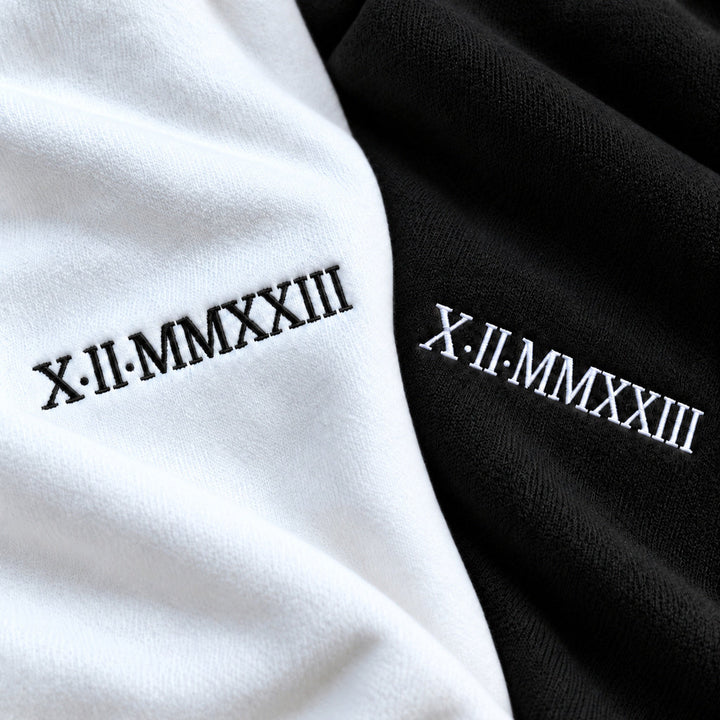 Macro de la date X.II.MMXXIII brodée en noir sur le sweat blanc et en blanc sur le sweat noir, avec texture des fils visible.