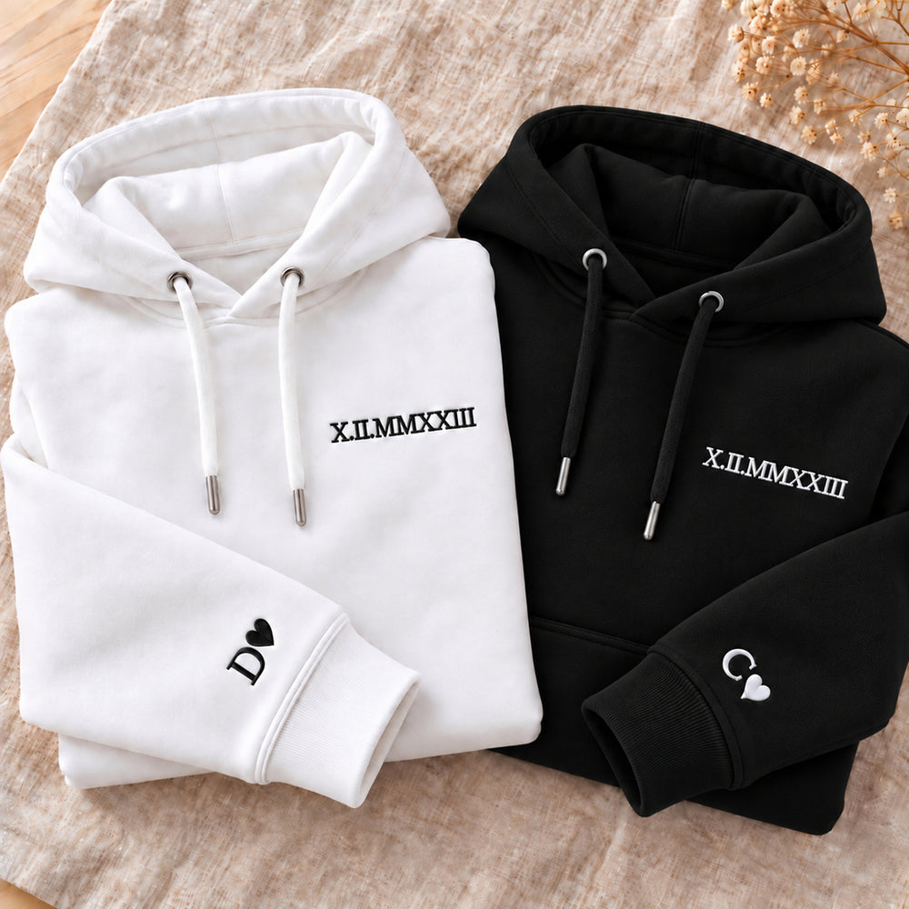 Deux sweats couple à capuche pliés, blanc à broderie noire et noir à broderie blanche, avec date X.II.MMXXIII et initiales D et C accompagnées de cœurs.