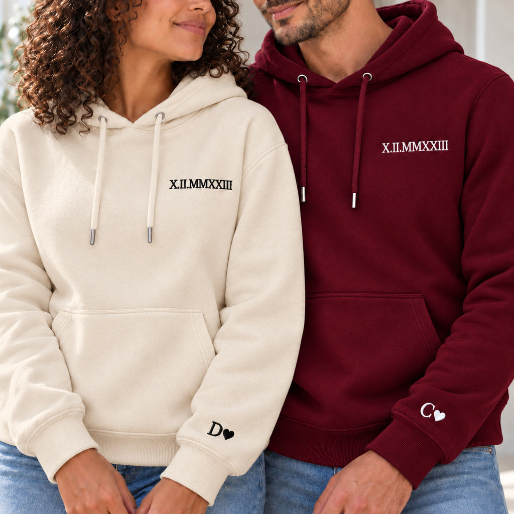 Couple en sweats personnalisés : femme à gauche en beige et homme en bordeaux, date en chiffres romains à la poitrine et initiales avec cœurs aux poignets.
