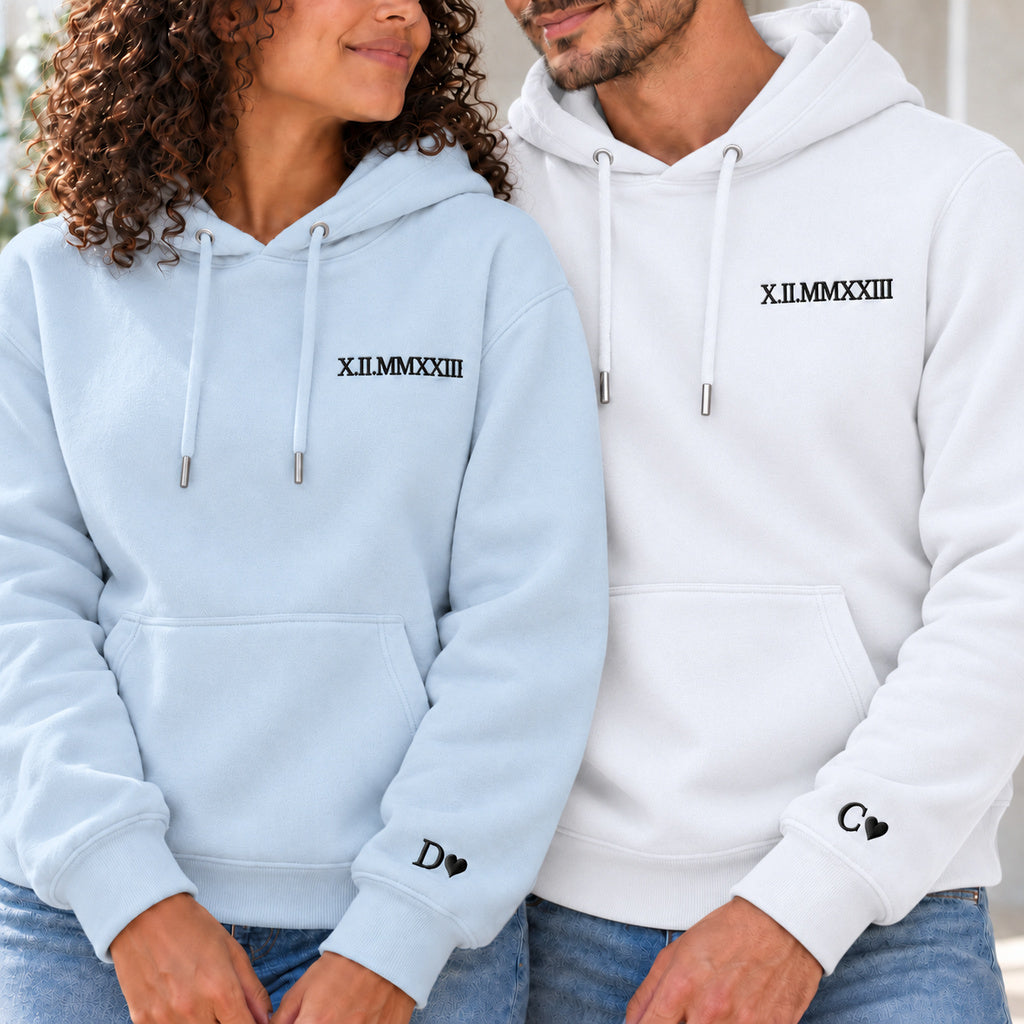 Couple en sweats personnalisés : femme à gauche en bleu et homme en blanc, date en chiffres romains à la poitrine et initiales avec cœurs aux poignets.