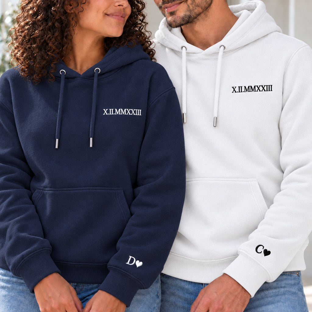 Couple en sweats personnalisés : femme à gauche en bleu marine et homme en blanc, date en chiffres romains à la poitrine et initiales avec cœurs aux poignets.