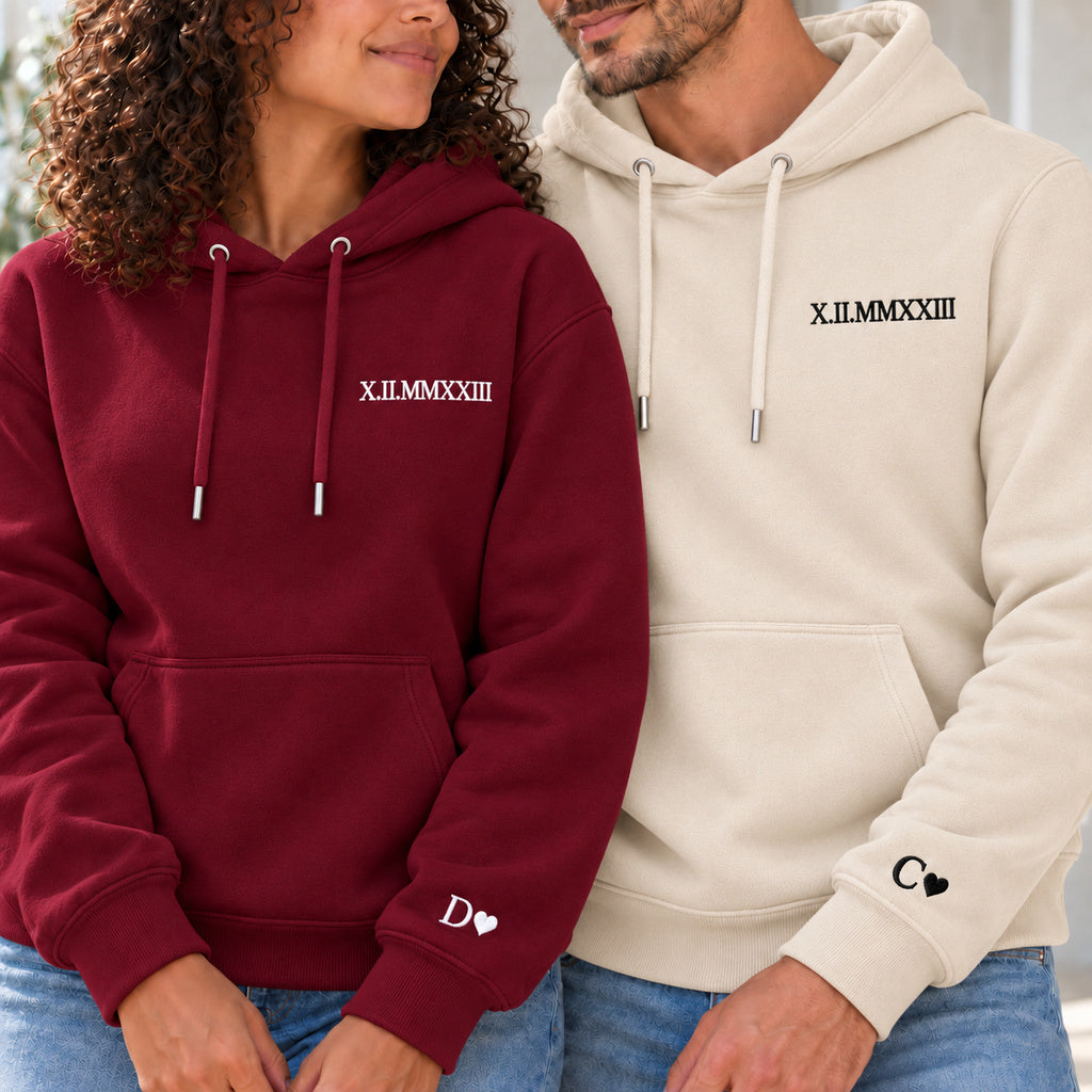 Couple en sweats personnalisés : femme à gauche en bordeaux et homme en beige, date en chiffres romains à la poitrine et initiales avec cœurs aux poignets.