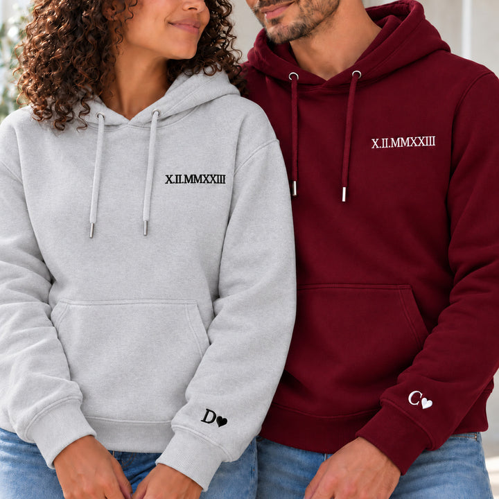 Couple en sweats personnalisés : femme à gauche en gris et homme en bordeaux, date en chiffres romains à la poitrine et initiales avec cœurs aux poignets.