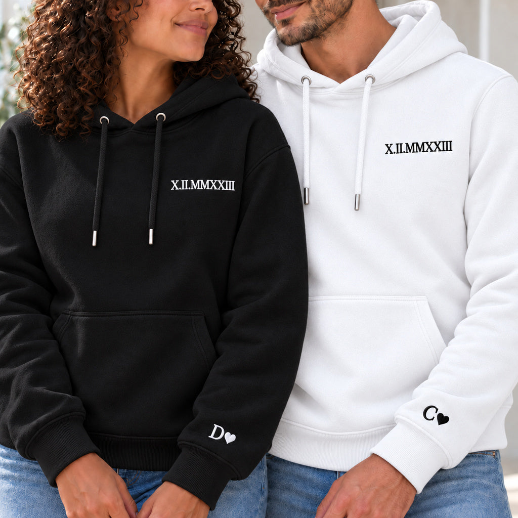 Couple en sweats personnalisés : femme à gauche en noir et homme en blanc, date en chiffres romains à la poitrine et initiales avec cœurs aux poignets.