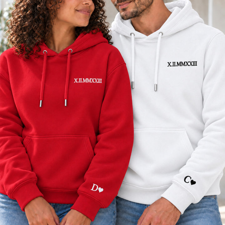 Couple en sweats personnalisés : femme à gauche en rouge et homme en blanc, date en chiffres romains à la poitrine et initiales avec cœurs aux poignets.