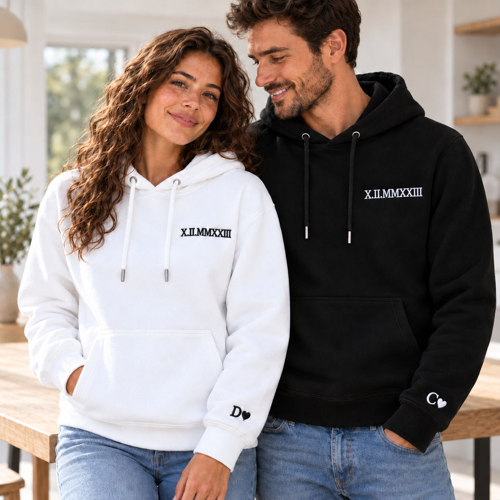 Couple souriant en pulls à capuche blanc et noir personnalisés avec la date X.II.MMXXIII et les initiales D et C accompagnées de cœurs aux poignets.