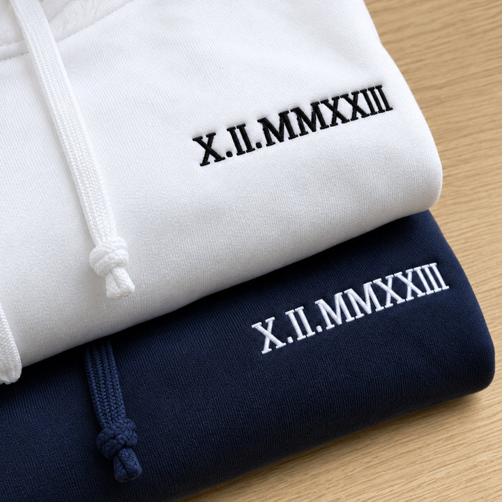 Gros plan de deux sweats pliés blanc et bleu marine, avec la date X.II.MMXXIII brodée en fils contrastants et des cordons noués.