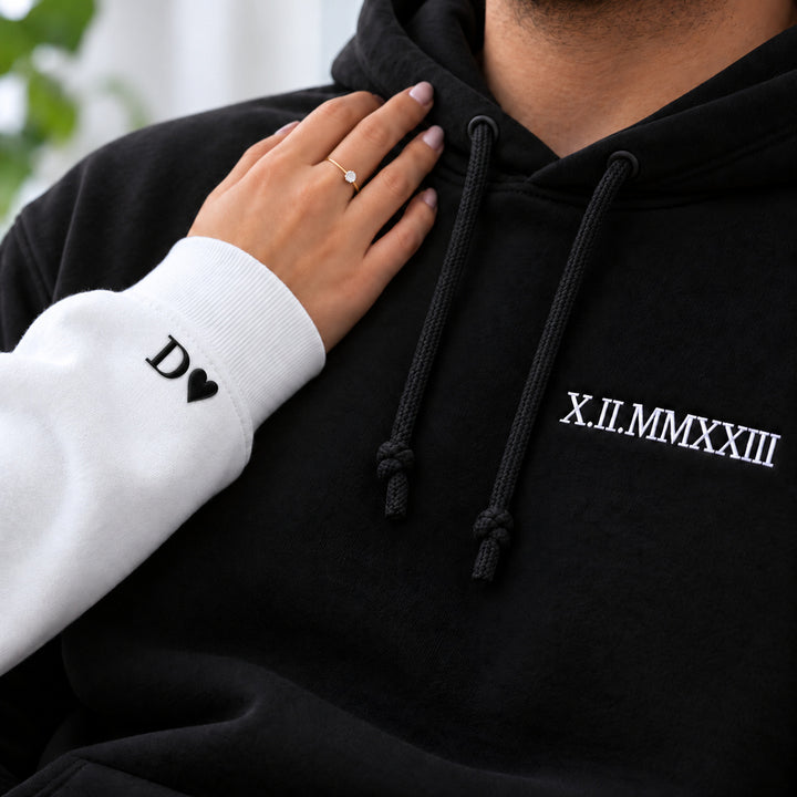 Main posée sur un sweat noir à date brodée blanche, avec initiale D et cœur noirs sur le poignet du sweat blanc, bague et cordons noués visibles.
