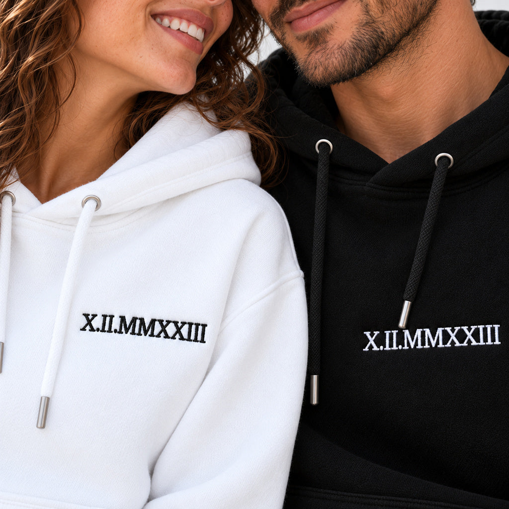 Plan rapproché des dates X.II.MMXXIII brodées en noir sur le pull blanc et en blanc sur le pull noir, portés par un couple.