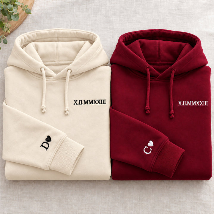 Deux sweats pliés : beige à gauche et bordeaux à droite, dates brodées et initiales D et C avec cœurs aux poignets, cordons noués.
