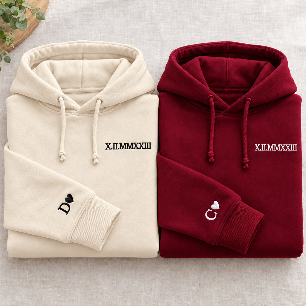 Deux sweats pliés : beige à gauche et bordeaux à droite, dates brodées et initiales D et C avec cœurs aux poignets, cordons noués.