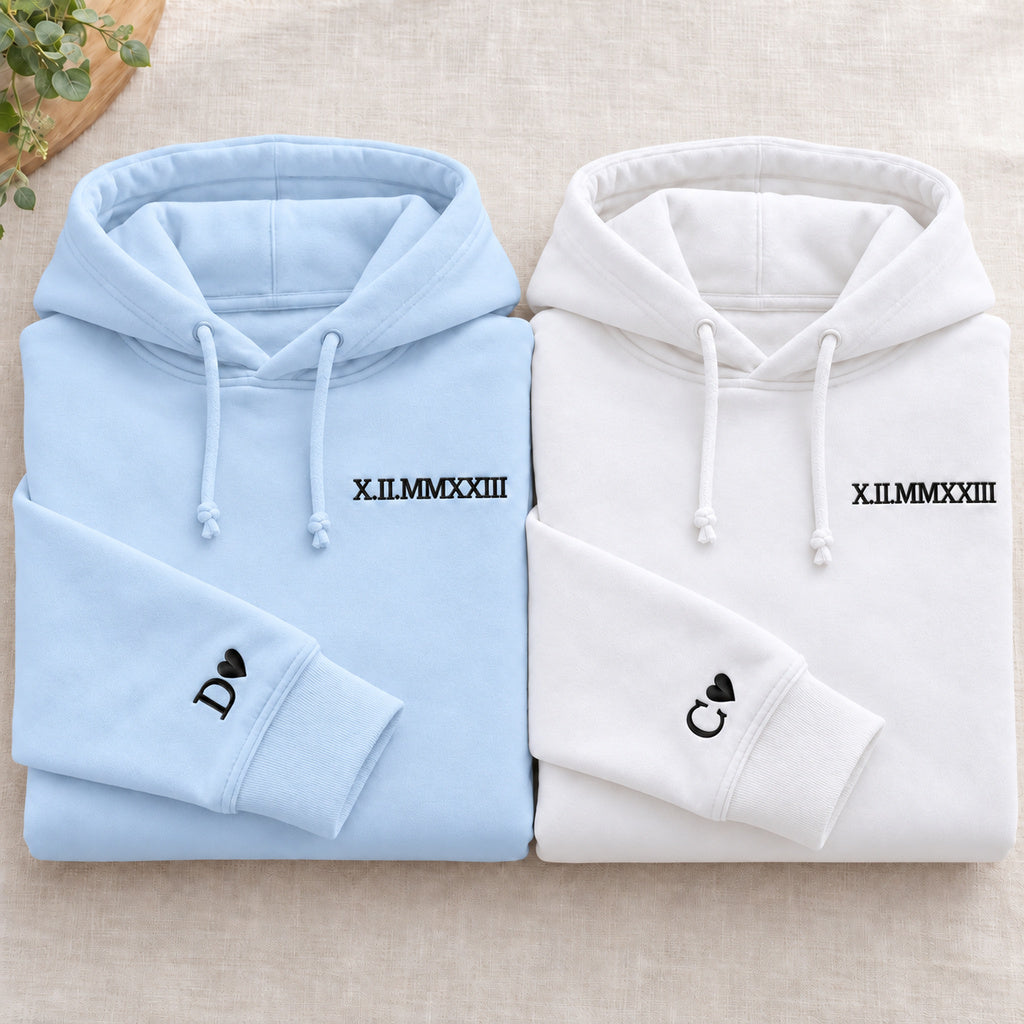 Deux sweats pliés : bleu à gauche et blanc à droite, dates brodées et initiales D et C avec cœurs aux poignets, cordons noués.