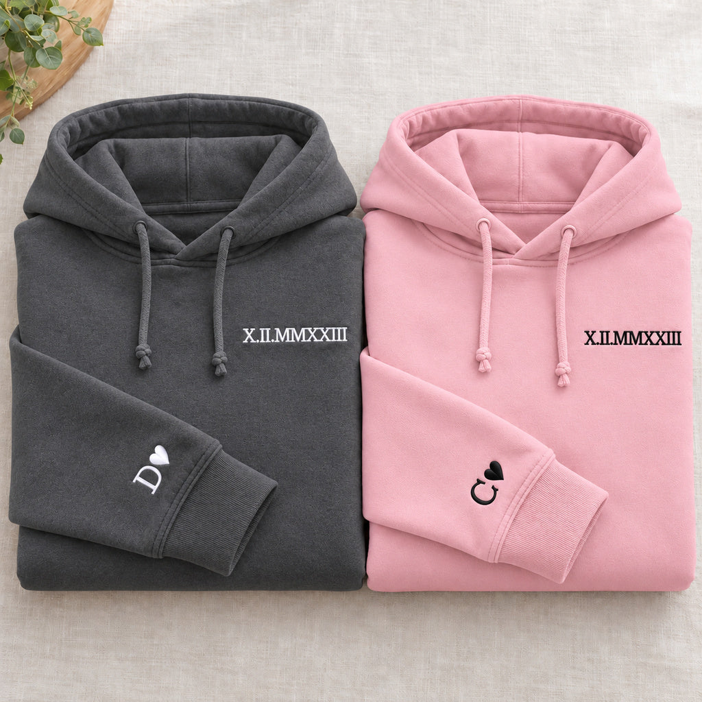 Deux sweats pliés : gris foncé à gauche et rose à droite, dates brodées et initiales D et C avec cœurs aux poignets, cordons noués.