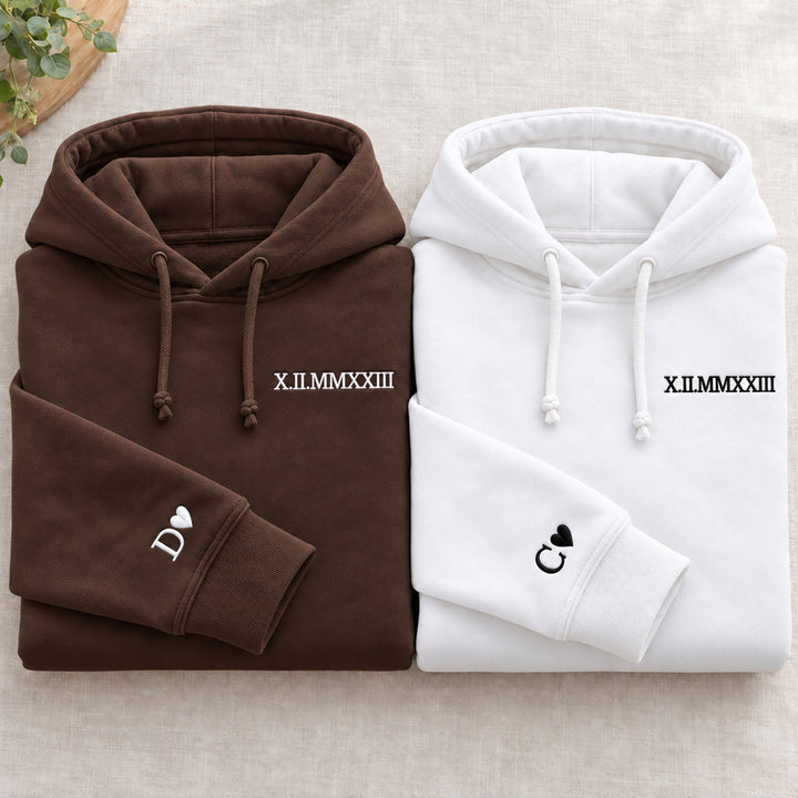 Deux sweats pliés : marron à gauche et blanc à droite, dates brodées et initiales D et C avec cœurs aux poignets, cordons noués.