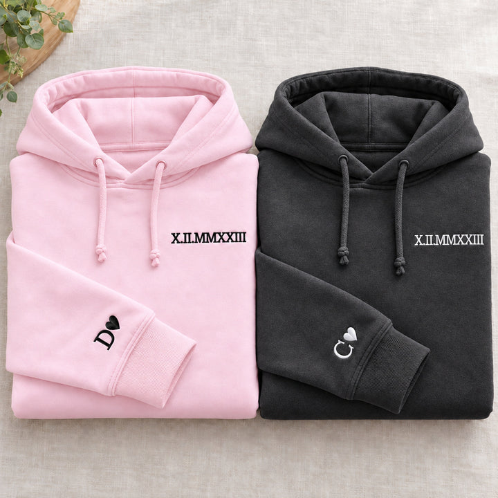 Deux sweats pliés : rose à gauche et gris foncé à droite, dates brodées et initiales D et C avec cœurs aux poignets, cordons noués.