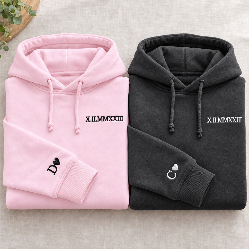 Deux sweats pliés : rose à gauche et gris foncé à droite, dates brodées et initiales D et C avec cœurs aux poignets, cordons noués.