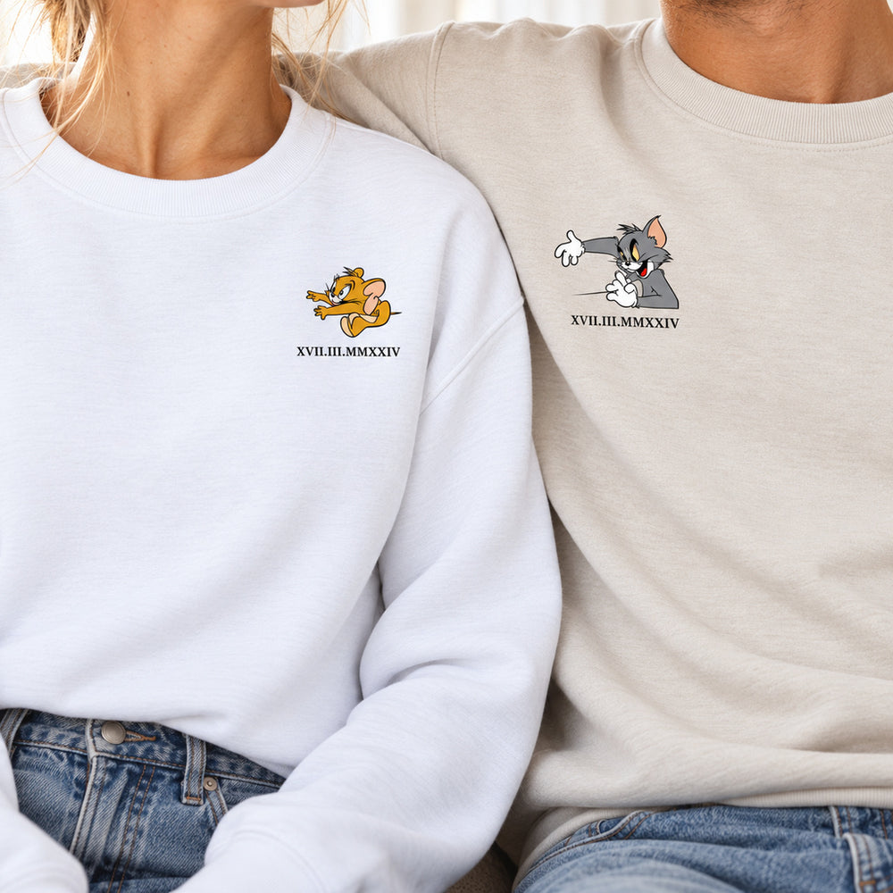 Nahaufnahme des Paares in weißen und beigen Sweatshirts mit Fokus auf die Jerry- und Tom-Aufdrucke und das personalisierte Datum in römischen Zahlen.