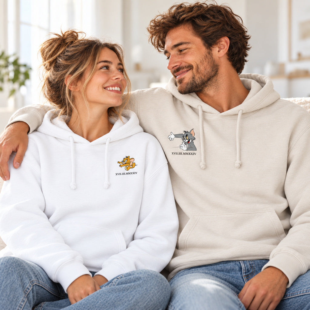 Sitzendes Paar in weißen und beigen Kapuzenpullovern mit Jerry- und Tom-Aufdrucken und personalisiertem Datum in römischen Zahlen.