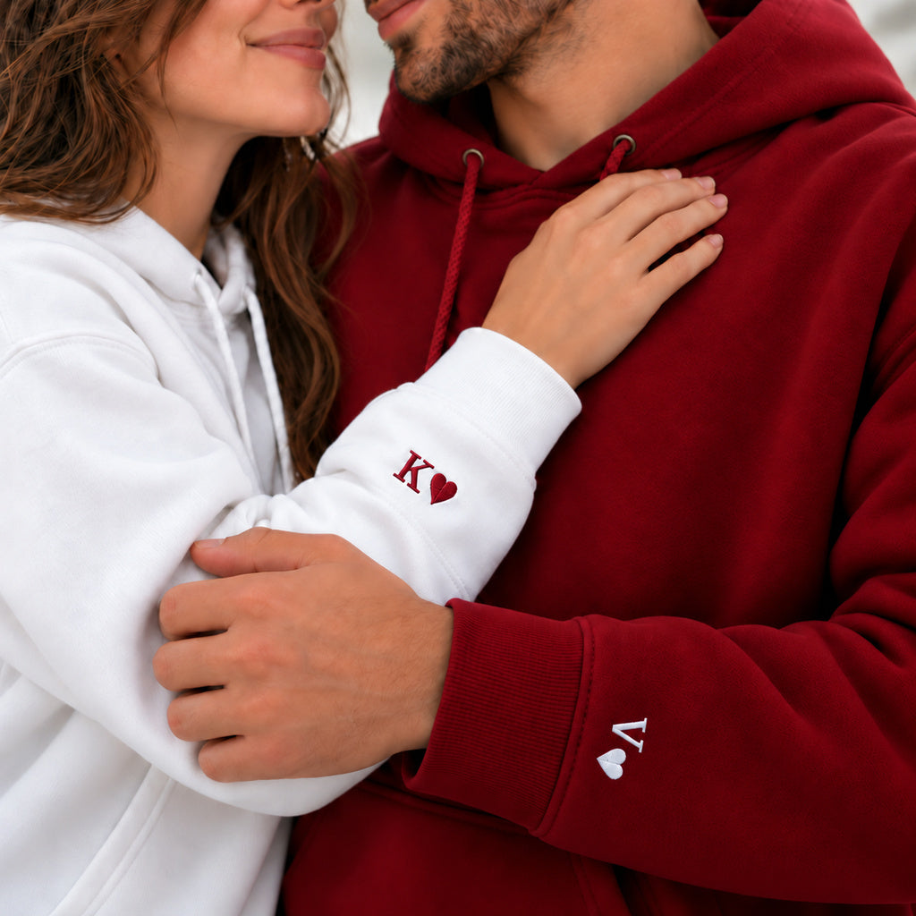Couple en sweats à capuche blanc et bordeaux, détail des initiales K et V et des petits cœurs brodés près des poignets.