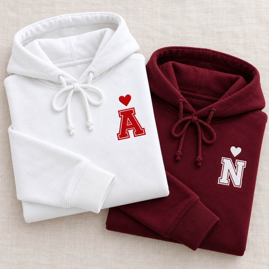 Deux sweats à capuche personnalisés pliés, blanc avec initiale A rouge et bordeaux avec initiale N blanche, chacun surmonté d’un cœur brodé.
