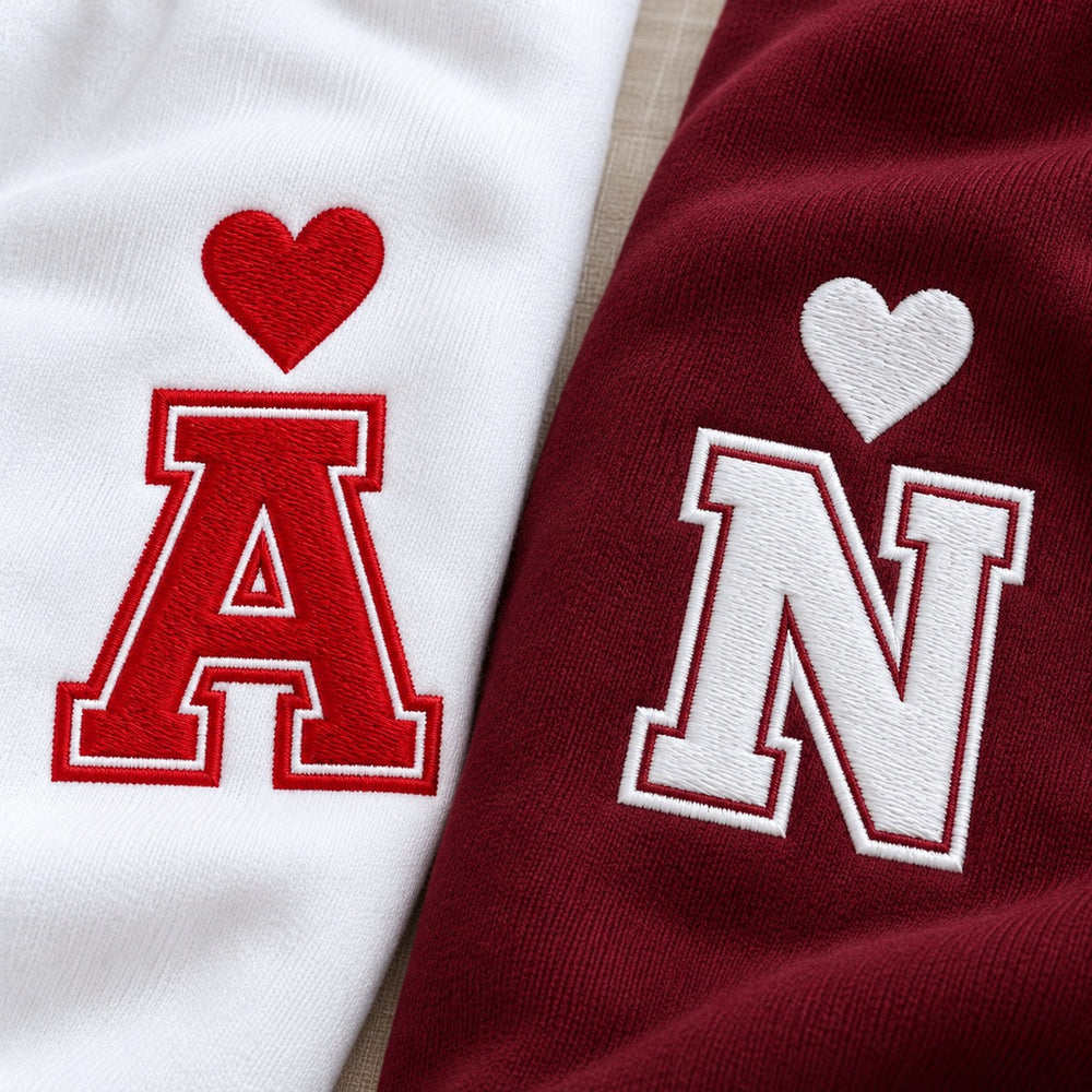 Gros plan des initiales A rouge sur sweat blanc et N blanche sur sweat bordeaux, avec cœurs et contours brodés style varsity.