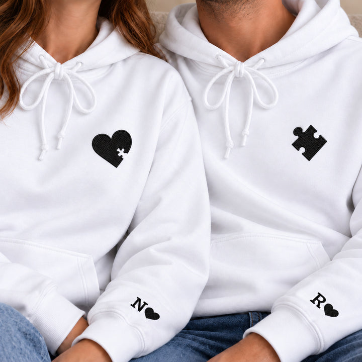 Gros plan des sweats blancs du couple : cœur et puzzle brodés noirs à la poitrine, N et R avec petits cœurs noirs aux poignets.
