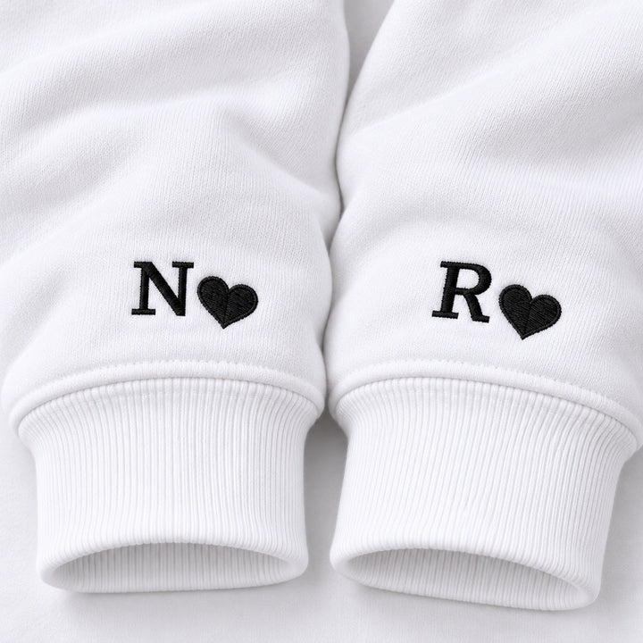 Macro des poignets des sweats blancs : initiale N et petit cœur noirs pour elle, initiale R et petit cœur noirs pour lui.