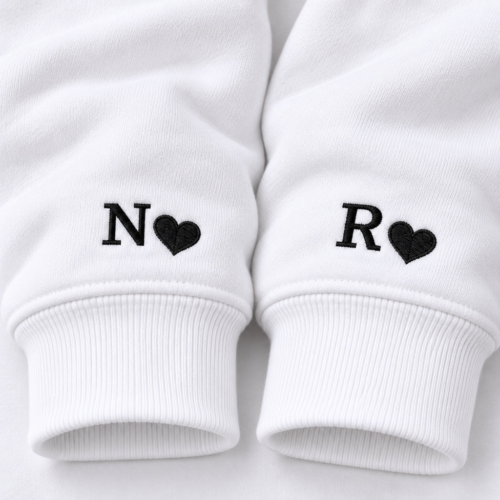 Macro des poignets des sweats blancs : initiale N et petit cœur noirs pour elle, initiale R et petit cœur noirs pour lui.