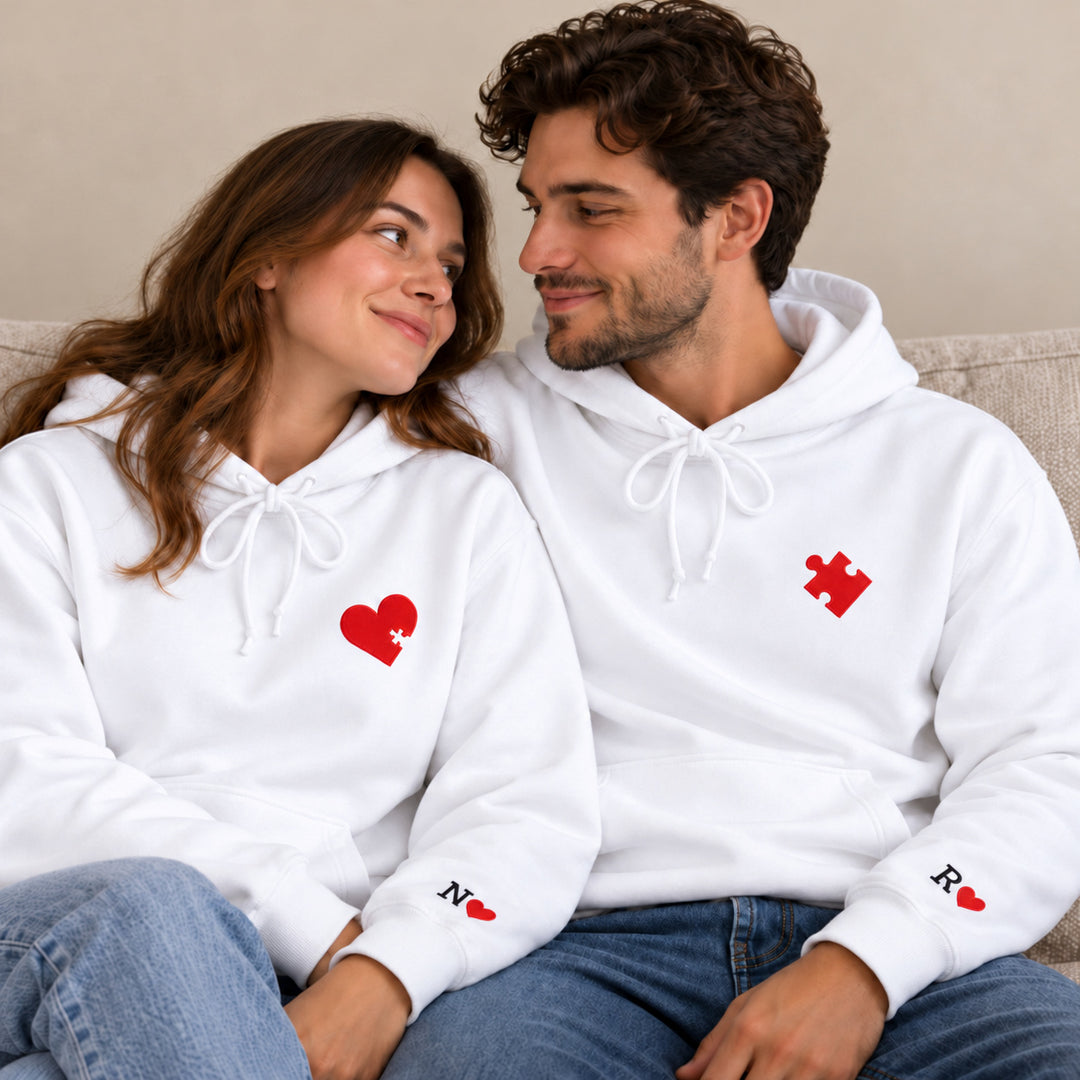 Sweats personnalisés couple blancs, cœur puzzle rouge et initiales N et R aux poignets, portés sur un canapé.