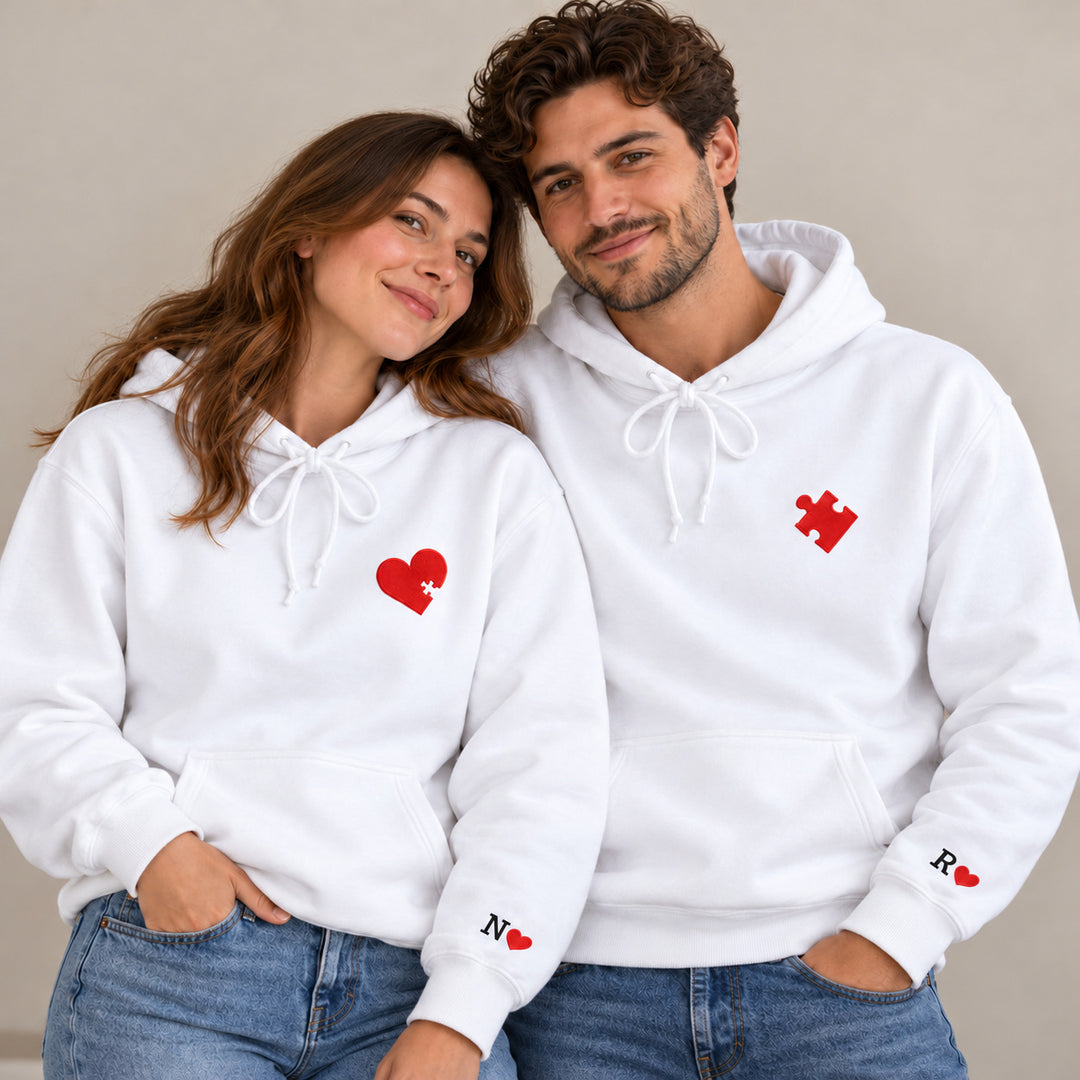 Couple debout en sweats blancs à capuche, cœur et pièce de puzzle rouges avec initiales N et R aux poignets.