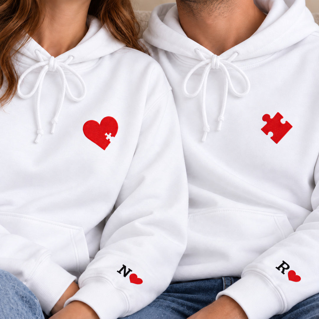 Gros plan des motifs cœur et pièce de puzzle rouges sur les sweats blancs, avec initiales N et R aux manches.
