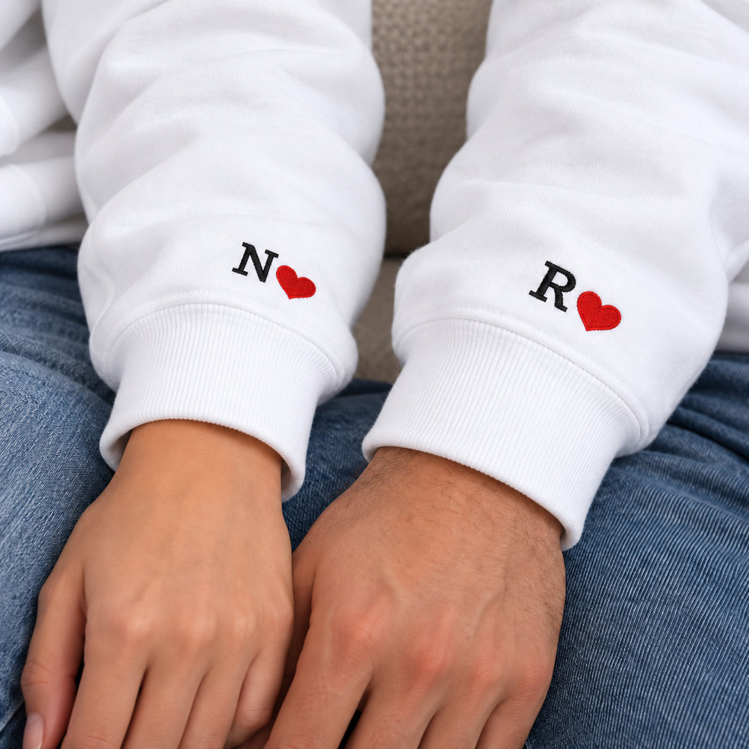 Détail des poignets des sweats blancs avec initiales brodées N pour la femme et R pour l’homme, accompagnées de cœurs rouges.